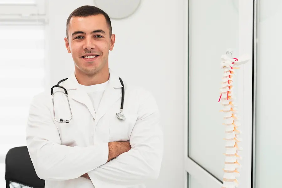 Médico ortopedista com estetoscópio ao lado de modelo da coluna vertebral, representando áreas de atuação da ortopedia e cuidado com o sistema musculoesquelético