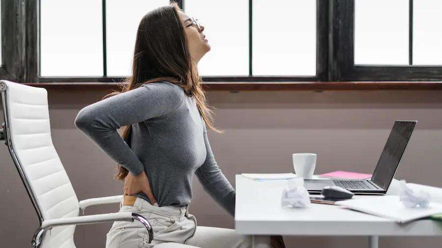 Mulher sentada em escritório segurando a região lombar enquanto trabalha no computador, ilustrando dores na lombar causadas por má postura.