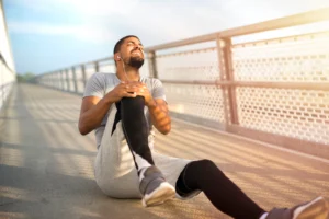 Pessoa sentada após corrida segura o joelho com expressão de dor, ilustrando desconforto muscular e articular após atividade física intensa.