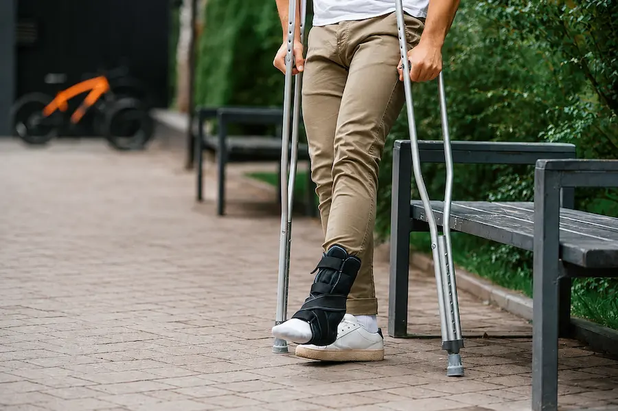 Pessoa usando bota ortopédica e muletas para caminhar em um parque.