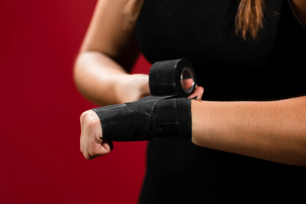 Mulher usando munhequeiras pretas durante exercício com halteres, em frente a um fundo vermelho.