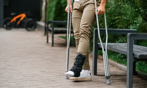 Pessoa usando bota ortopédica e muletas para caminhar em um parque.