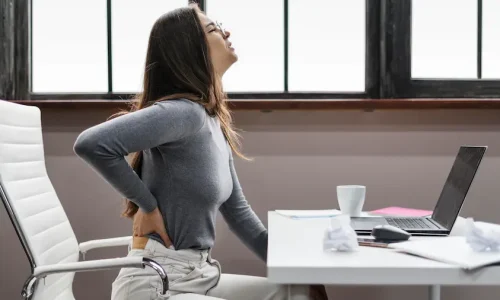 Mulher sentada em escritório segurando a região lombar enquanto trabalha no computador, ilustrando dores na lombar causadas por má postura.