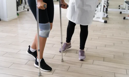 Paciente utilizando muletas e joelheira durante sessão de fisioterapia para recuperação de fratura por estresse.