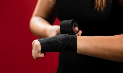 Mulher usando munhequeiras pretas durante exercício com halteres, em frente a um fundo vermelho.